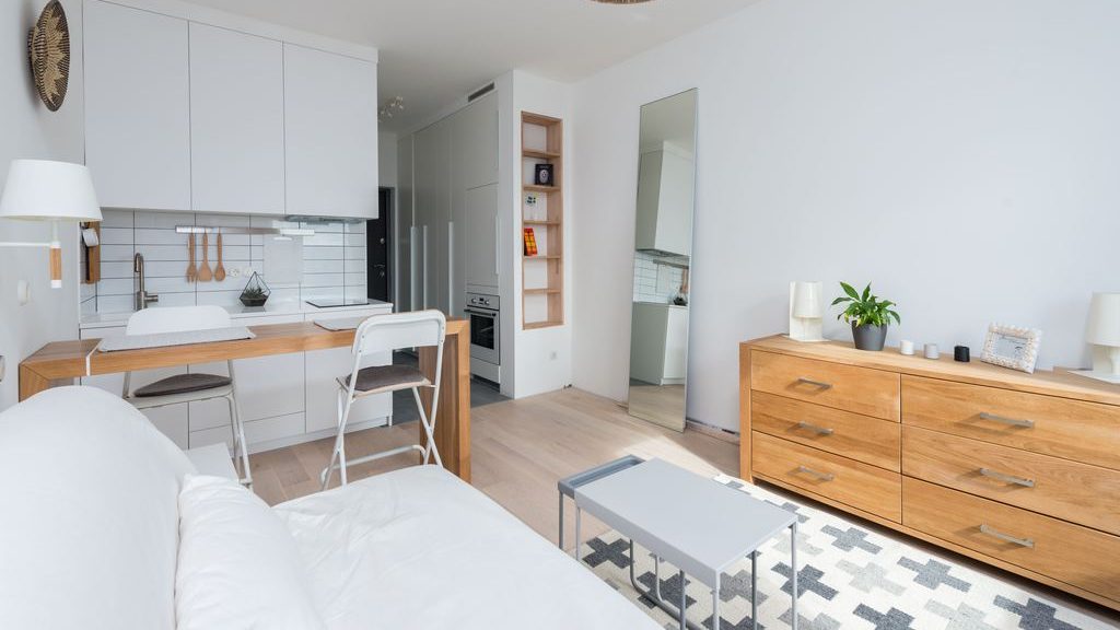 Compact tiny home interior with white sofa, small kitchen, wood dresser, coffee table, mirror, and light neutral decor