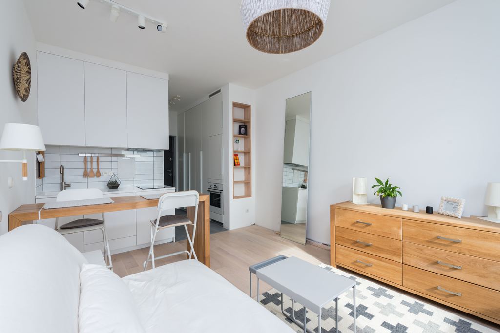 Bright studio apartment with a white sofa, compact kitchen, foldable dining table, wooden dresser, and floor mirror.