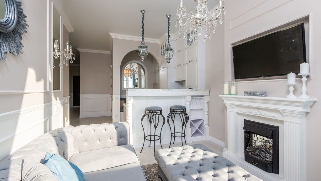Wall-mounted TV above a white fireplace in an elegant living room with a tufted sectional sofa, ottoman, crystal chandeliers, and an open kitchen bar.