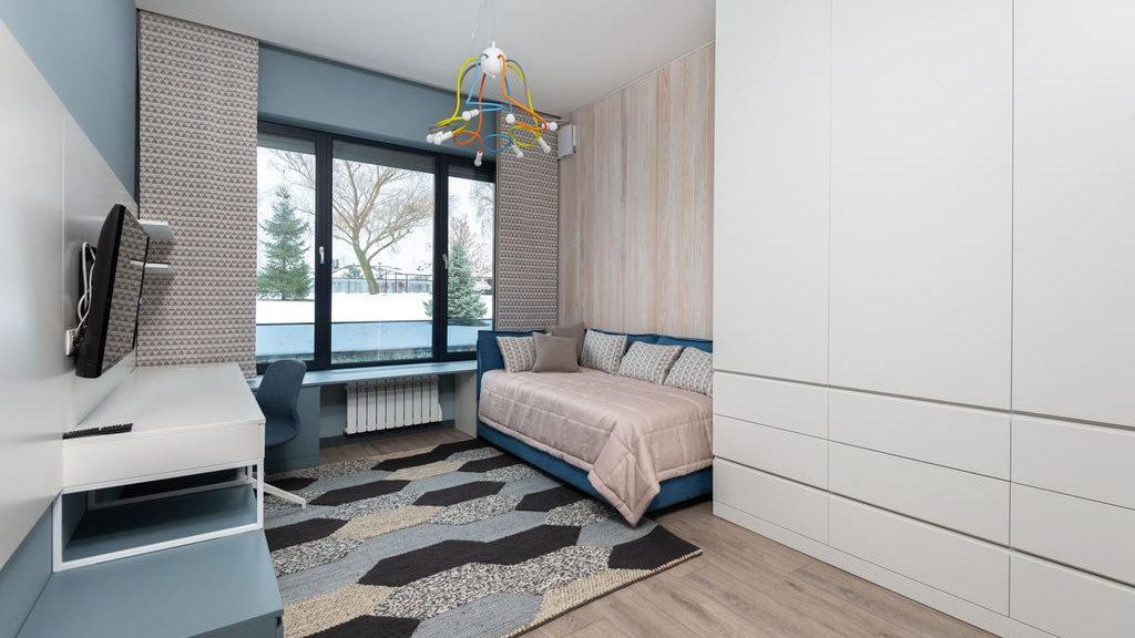 Blue upholstered daybed in a compact multipurpose room with a desk, wall-mounted TV, and built-in wardrobe.