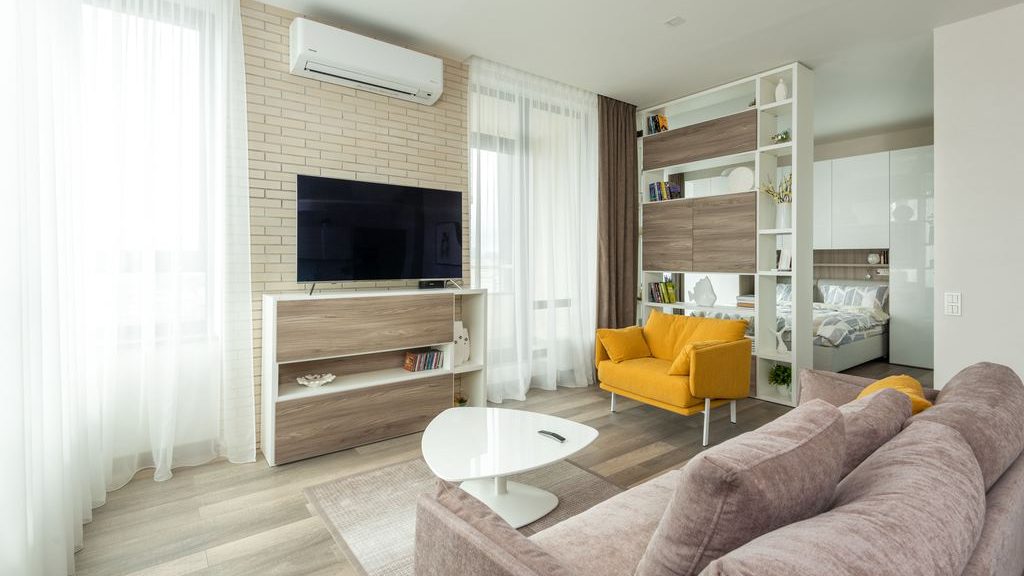 Modern studio apartment with a sofa, yellow accent chair, TV console, open shelving divider, and a bed in the background.