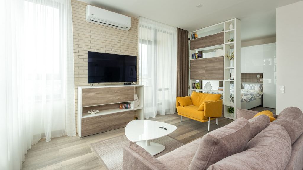 Open-plan studio living room with a TV console, white shelving divider, yellow armchair, sofa, and bedroom area behind.