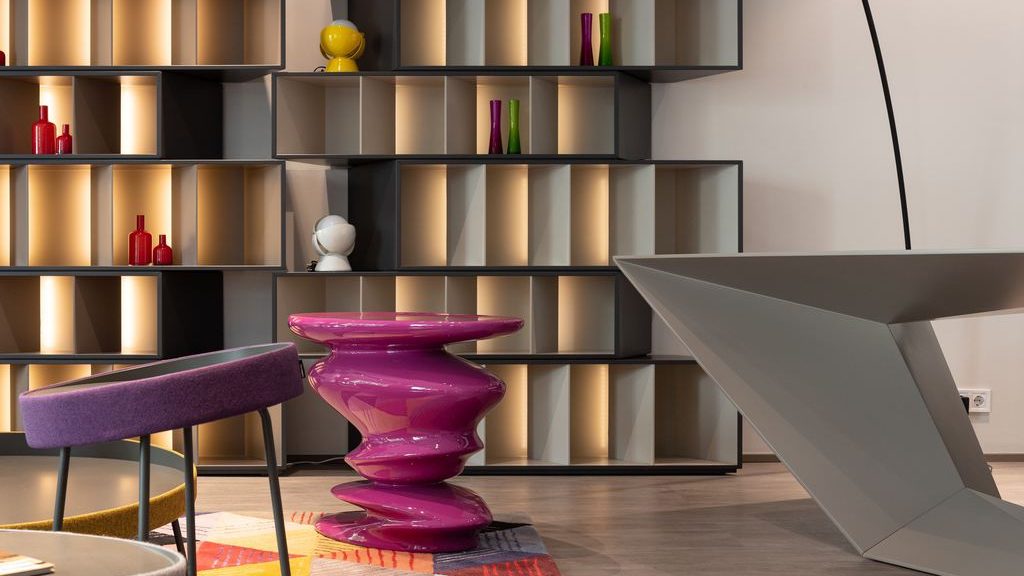 Postmodern interior with illuminated geometric shelving, a glossy purple sculptural table, colorful rug, and angular gray furniture.