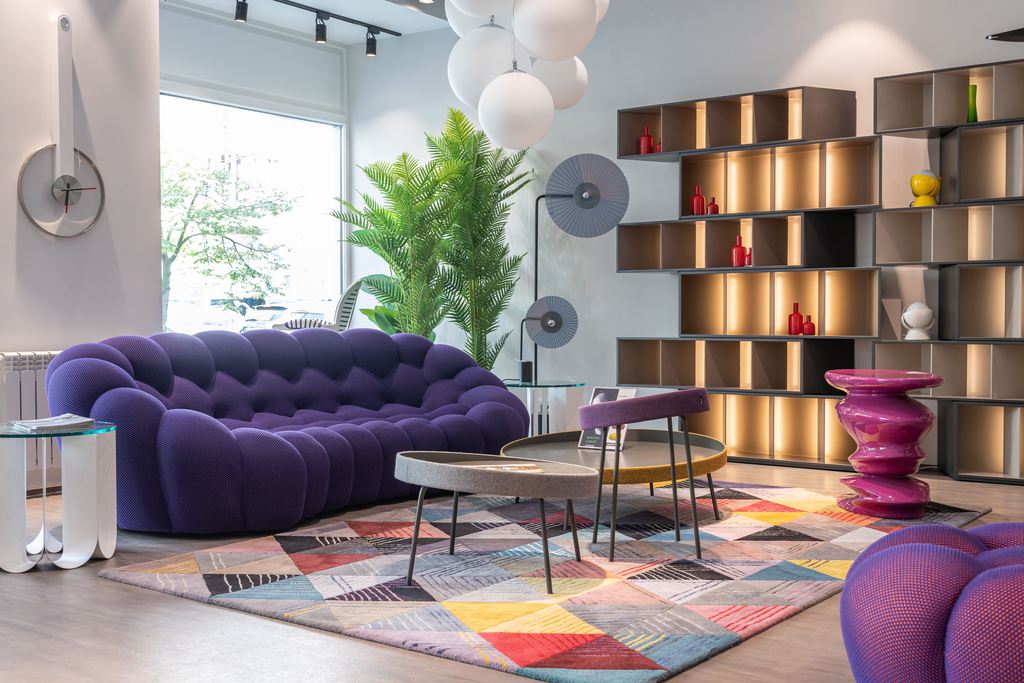 Postmodern living room with a curved purple sofa, geometric rug, sculptural tables, and illuminated modular shelving.