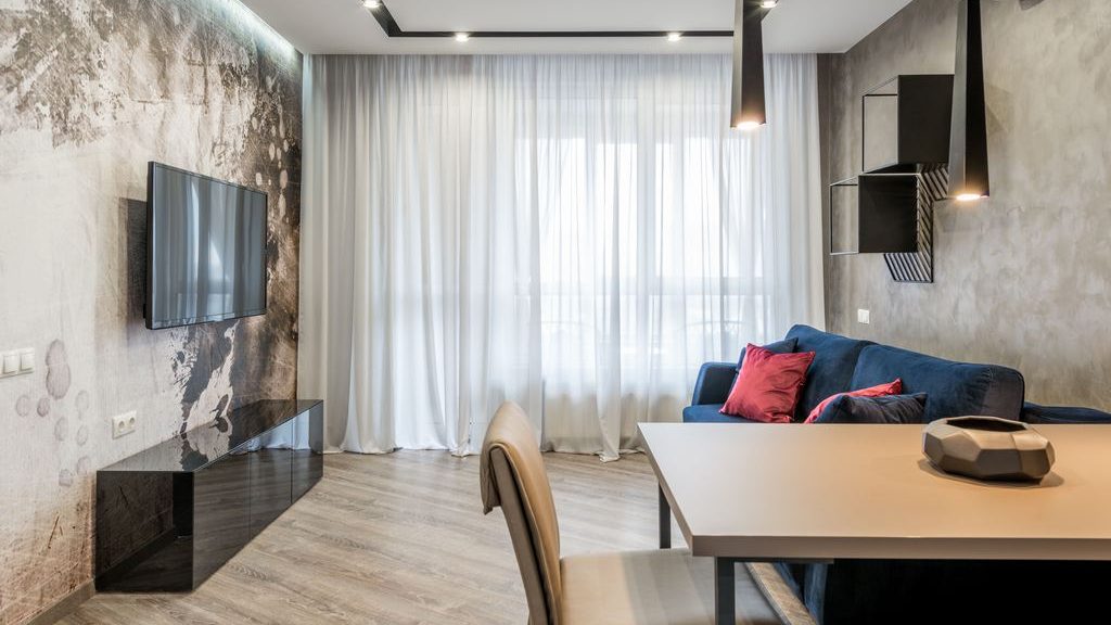 Wall-mounted TV above a glossy black media console in a modern gray living room with a blue sofa, sheer curtains, pendant lights, and a dining table.
