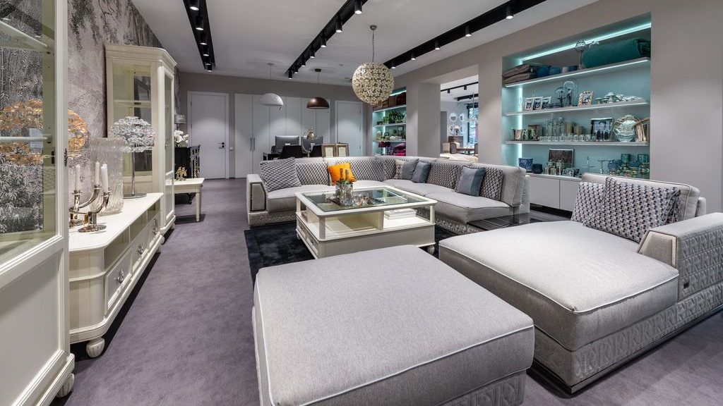 Large gray sectional sofa with patterned cushions, oversized ottoman, glass coffee table, built-in display shelving, and modern track lighting in an elegant living room.