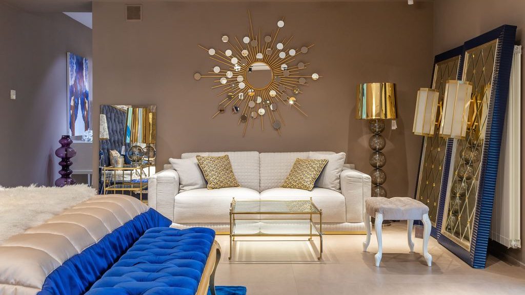 Modern glam living room with white sofa, blue velvet seating, gold coffee table, sunburst mirror, and reflective gold decor accents.
