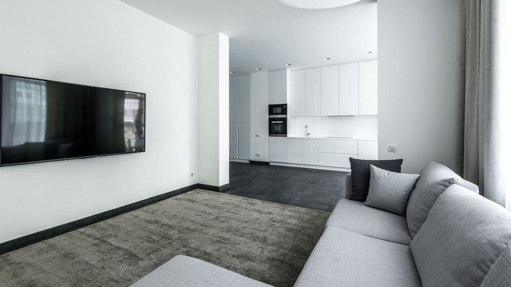 Wall-mounted TV in a bright minimalist living room with a gray sectional sofa, large area rug, floor-to-ceiling curtains, and an open white kitchen in the background.