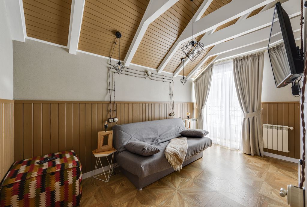 Gray sofa bed in a bright loft room with a wall-mounted TV, large window, wood-paneled walls, and open floor space.
