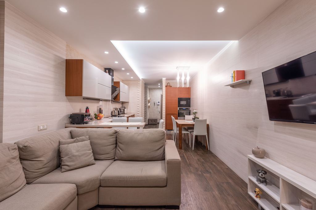 Bright studio apartment with a sectional sofa, open kitchen, dining table, wall-mounted TV, and built-in storage.