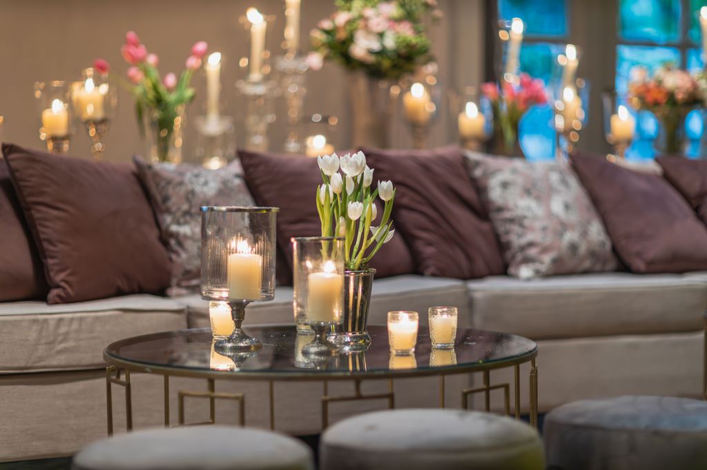 Lit candles and white tulips on a round coffee table in a cozy living room with a sofa and soft floral decor.