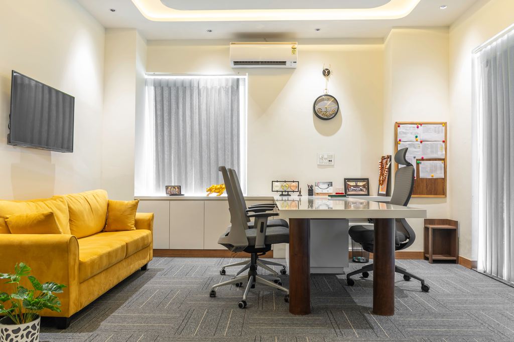 Modern office with a yellow sofa, conference table, ergonomic chairs, and wall-mounted TV.