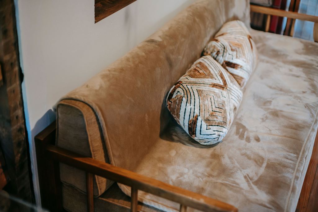 Beige futon sofa bed with a wooden frame and patterned bolster pillow in a cozy room.