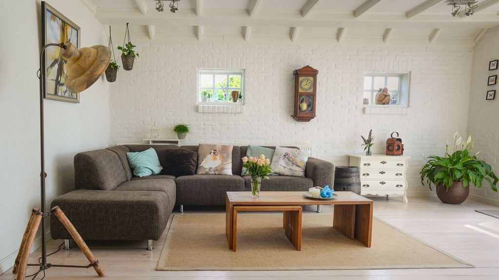 Dark gray sectional sofa with pastel pillows, wooden coffee tables, indoor plants, and white brick walls in a bright living room.
