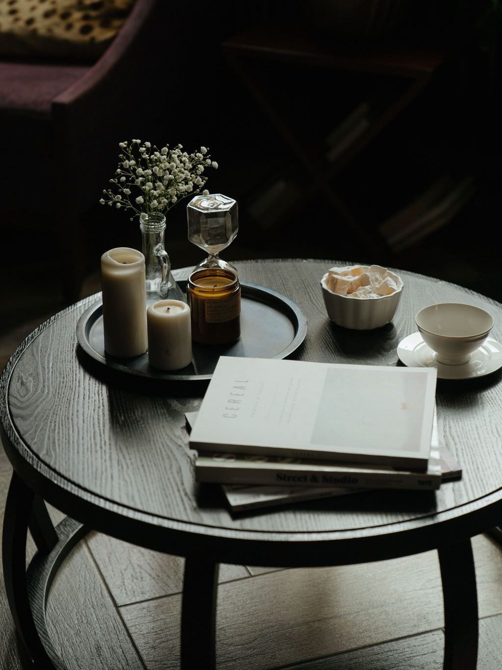 Dark wood round coffee table styled with candles, a floral vase, books, and a tea setting in a moody living room.