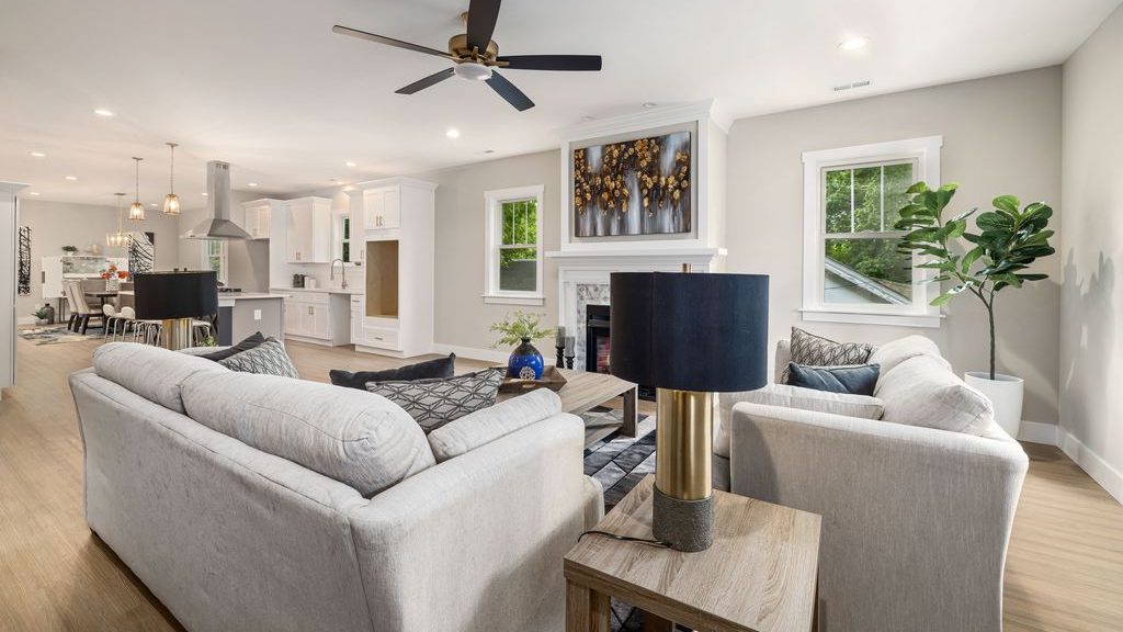 Open-concept living room with white walls, light gray sofas, a wood coffee table, dark accent lamps, and a bright white kitchen in the background.