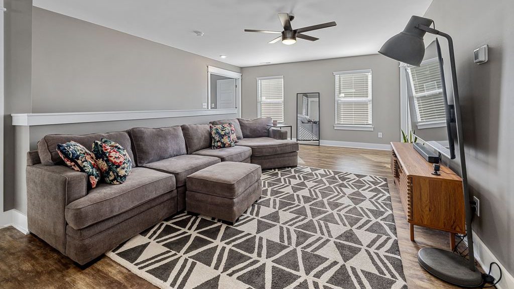 Gray sectional sofa with floral pillows and ottoman in a modern living room with a patterned rug, wood TV stand, and floor lamp.