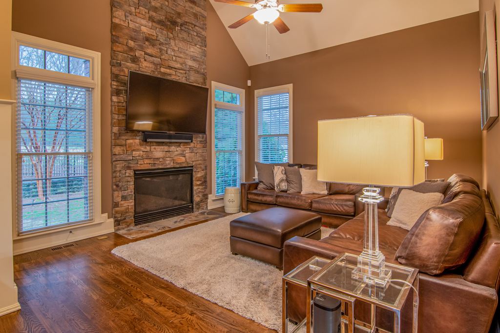 Brown leather sectional sofas in a living room with a stone fireplace, wall-mounted TV, beige rug, and dark wood flooring.