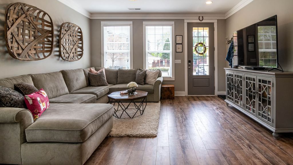 Bright living room with a gray sectional sofa, light rug, dark wood flooring, large windows, and a TV on a mirrored media console.