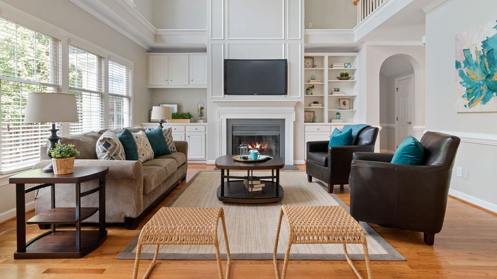 Bright transitional living room with a fireplace, gray sofa, dark leather chairs, teal accent pillows, and woven stools.