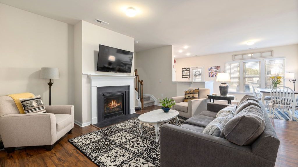 Bright open-plan living room with gray sofa, two accent chairs, marble coffee table, fireplace, patterned rug, and dining area.