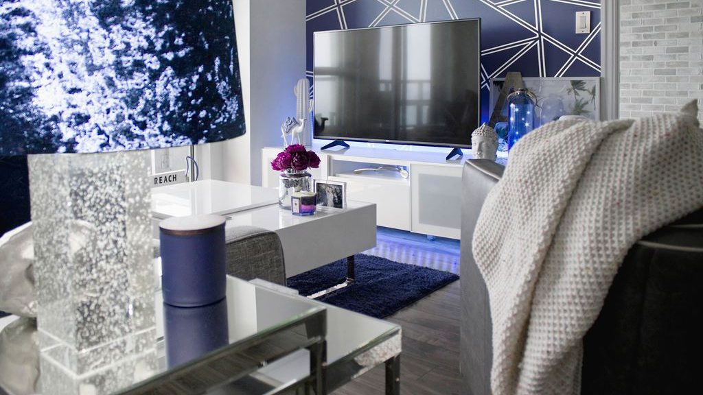 Modern living room with a geometric navy accent wall, white TV console, mirrored furniture, and blue ambient lighting.