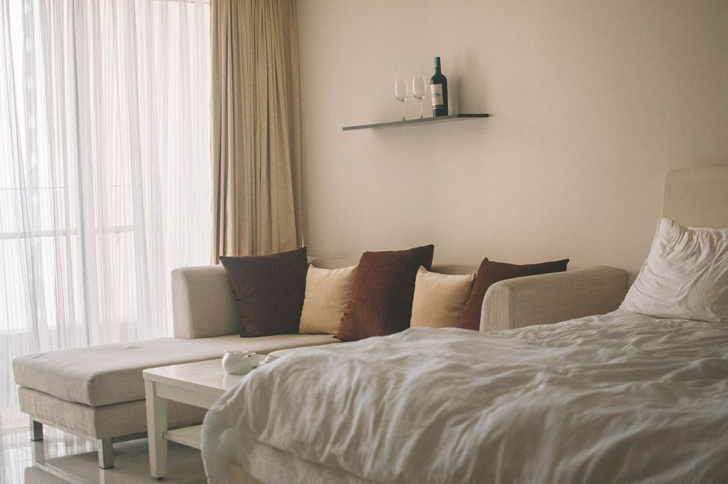 Beige sofa beside a made bed in a bright guest room with floor-to-ceiling curtains and a small coffee table.
