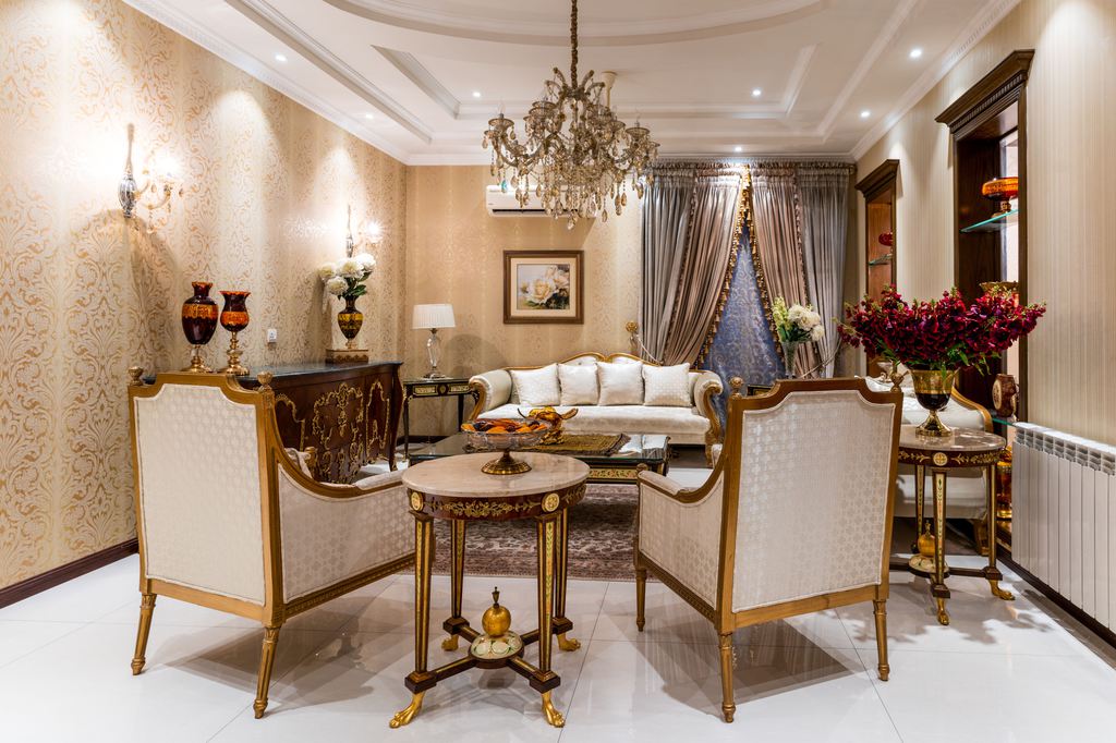 Classic luxury living room with gold-trimmed chairs, ornate tables, chandelier, patterned wallpaper, and elegant cream seating.