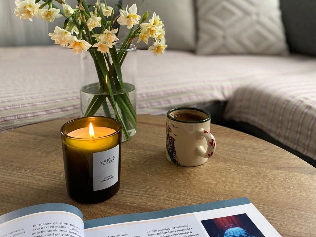 Lit candle, coffee mug, flowers, and an open magazine on a round wooden table in a cozy bedroom seating area.