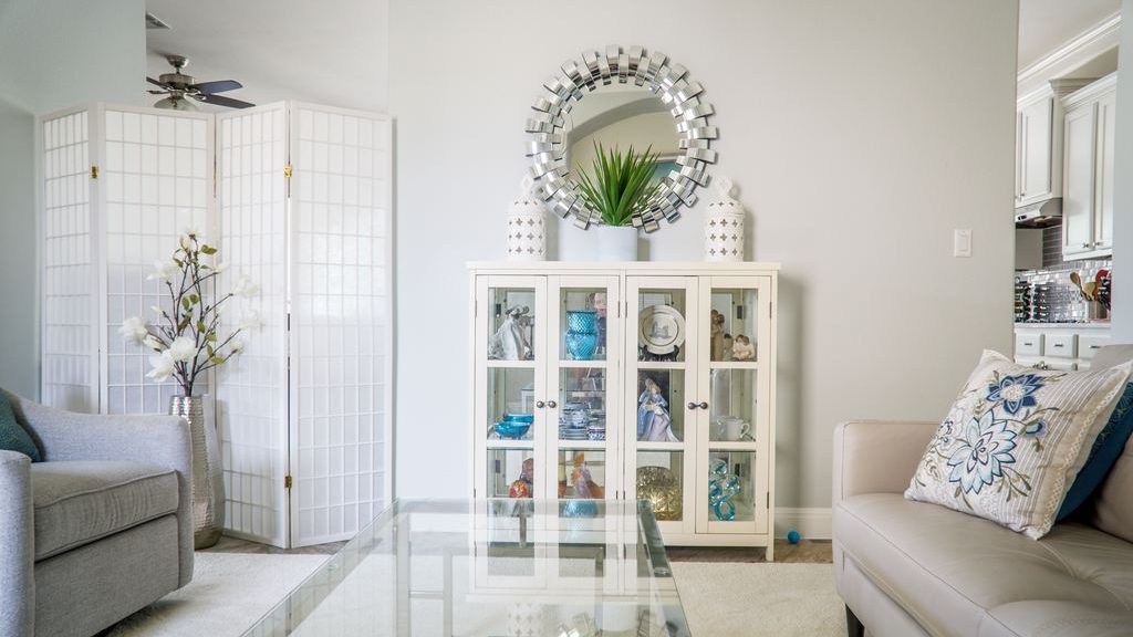 Bright living room with a round mirrored wall mirror above a white glass-front display cabinet, flanked by gray and cream seating.