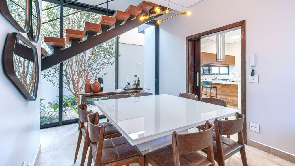 Modern dining room with a glossy square table, brown leather chairs, floor-to-ceiling windows, and a floating wood staircase.