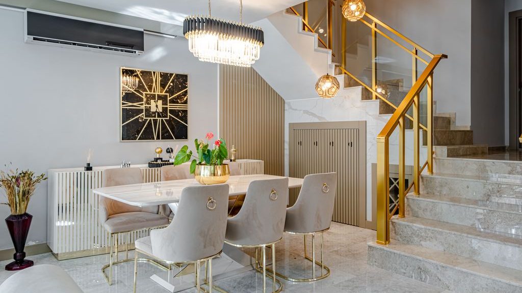 Luxury dining area with white marble table, cream chairs with gold legs, gold staircase railing, chandelier, and marble flooring.