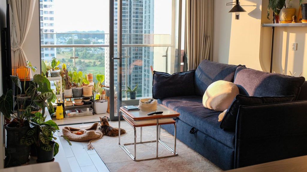 Cozy apartment living room with a dark blue sofa, indoor plants, a small coffee table, and city views through floor-to-ceiling balcony doors.