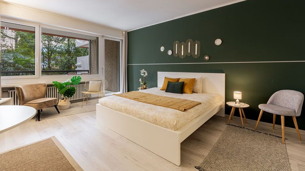 Studio apartment sleeping area with a white platform bed, dark green accent wall, two chairs, and large windows.