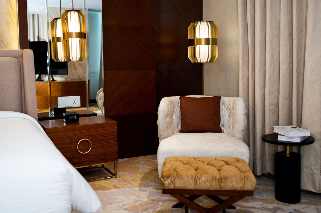 Cream upholstered accent chair with a brown pillow and matching ottoman beside a bed in a warm modern bedroom.