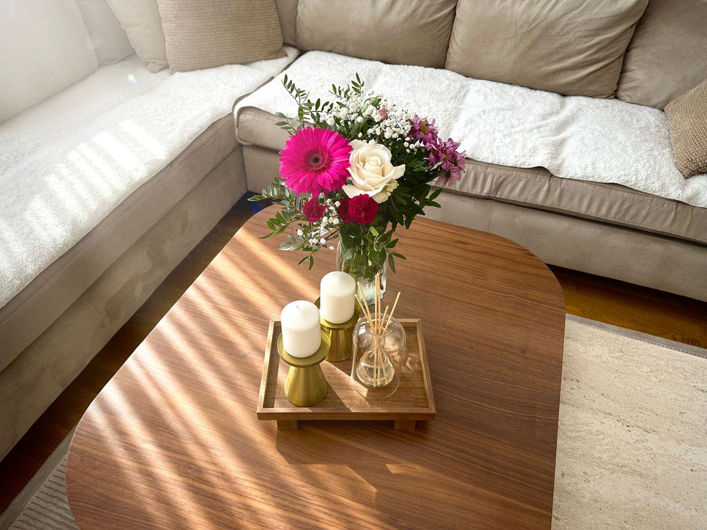 Colorful fresh flowers in a clear vase on a walnut coffee table with candles and a decorative tray beside a beige sectional sofa.