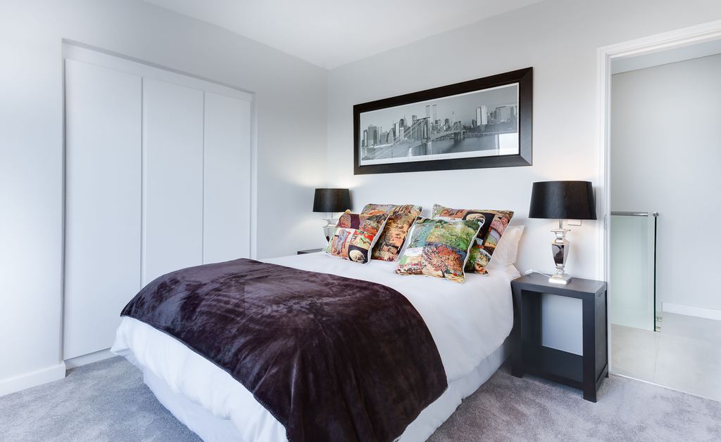 White bedroom with a dark throw blanket, black nightstands, colorful pillows, and a framed cityscape above the bed.