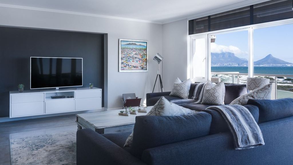 Modern living room with a dark blue sectional, white TV stand, framed wall art, and a black accent wall beside large ocean-view windows.