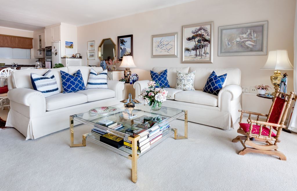 Bright living room with white sofas, blue patterned pillows, a glass coffee table with flowers and books, and a wooden rocking chair.