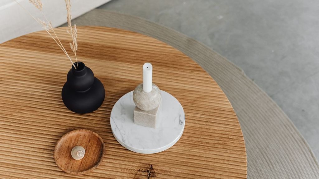 Minimal candle decor on a round wooden table with a black vase, dried stems, and a white candle on a marble tray.