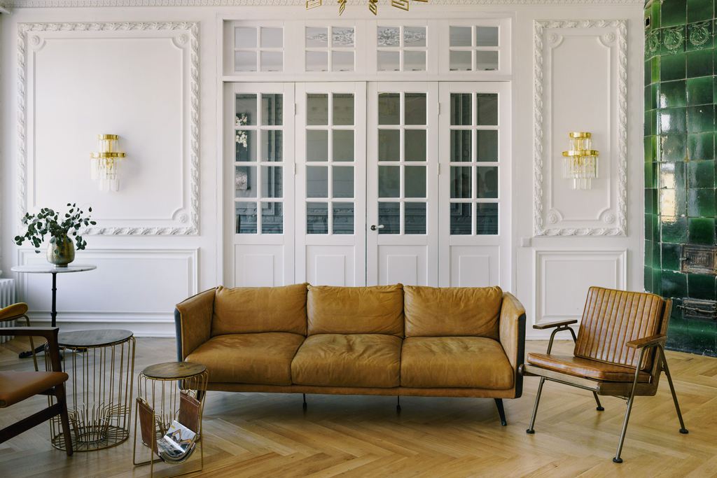 Cognac brown leather sofa with slim black legs in a bright living room with white paneled walls, herringbone wood flooring, and a matching leather chair.