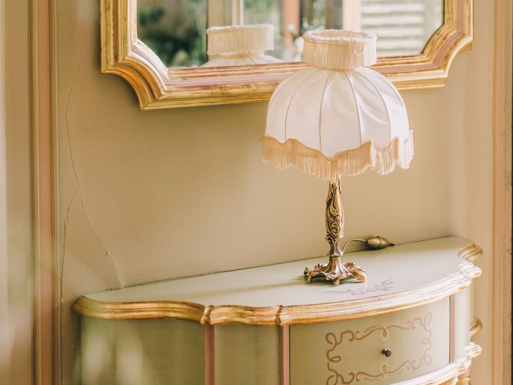 Vintage console table with gold trim, matching gold-framed mirror, fringed table lamp, and soft green wall decor in a classic interior.