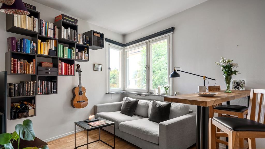 Tiny home living area with gray sofa, wall-mounted bookshelves, small coffee table, high dining table, and corner windows