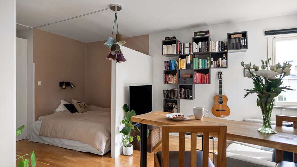 Studio apartment with a partially enclosed bed nook, dining table, wall-mounted bookshelves, and plants.