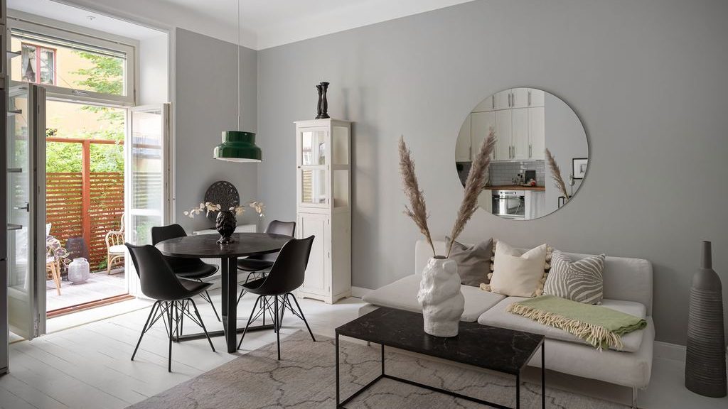 Bright open-plan studio with a white sofa, round wall mirror, black dining table, and glass doors to a small patio.