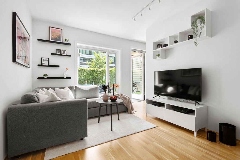 Alt text: Modern living room with white walls, gray sectional sofa, black coffee table, white TV stand, floating shelves, and light wood flooring.