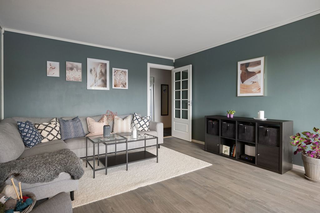 Gray sectional sofa with patterned pillows, framed wall art, and a black storage cabinet against deep teal living room walls.