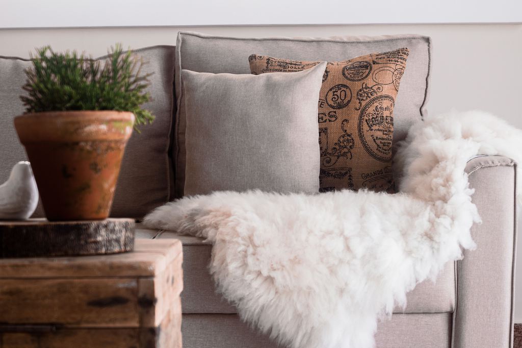 Light gray sofa styled with layered neutral and patterned pillows, a white faux-fur throw, rustic wood table, and potted plant.