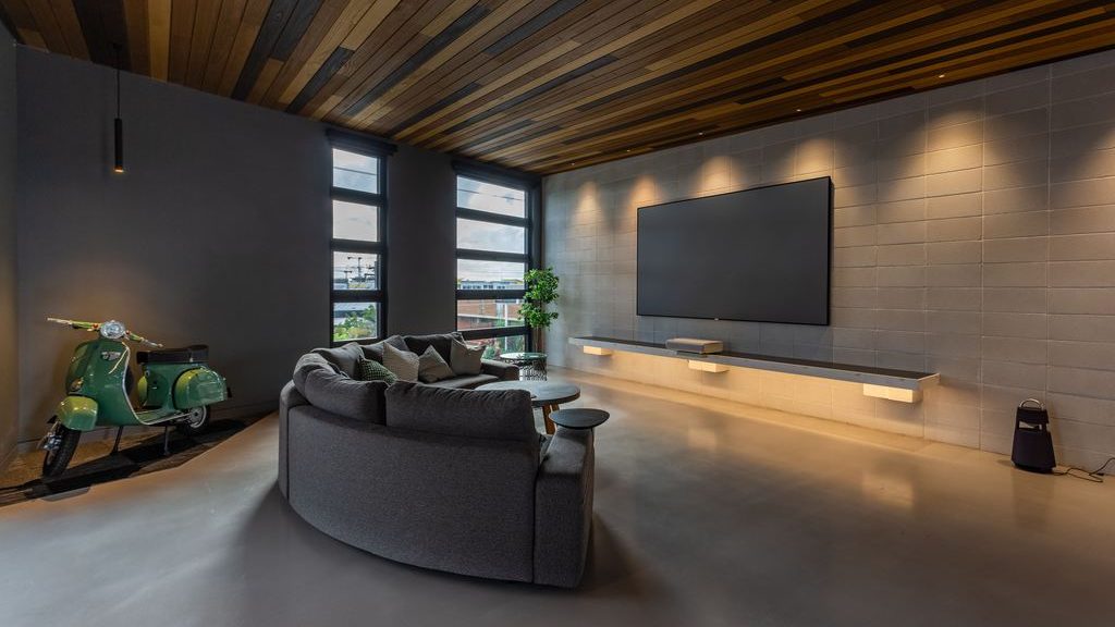 Modern home theater living room with a wall-mounted TV, curved sectional sofa, floating media shelves, and warm accent lighting.