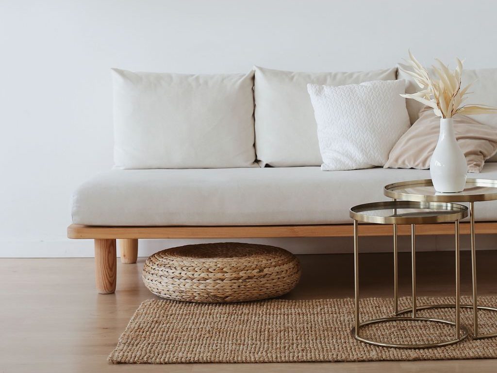 Minimal living room with a cream sofa, woven rug and floor cushion, brass nesting tables, and dried foliage in a white vase.
